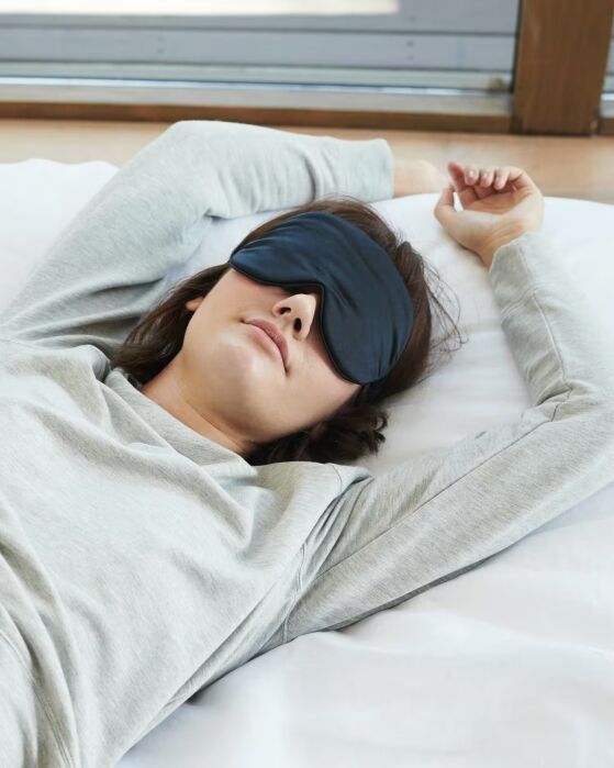 A person wearing a grey long-sleeve shirt and a black sleep mask lies on a bed with one arm resting above their head.
