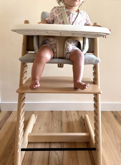 A baby wearing a floral outfit sits in a wooden high chair with feet dangling, tray attached, and a safety harness fastened.
