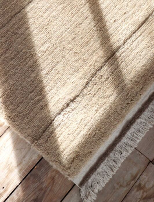 A beige textured rug with fringed edges lies on a wooden floor, with sunlight and shadows falling across its surface.