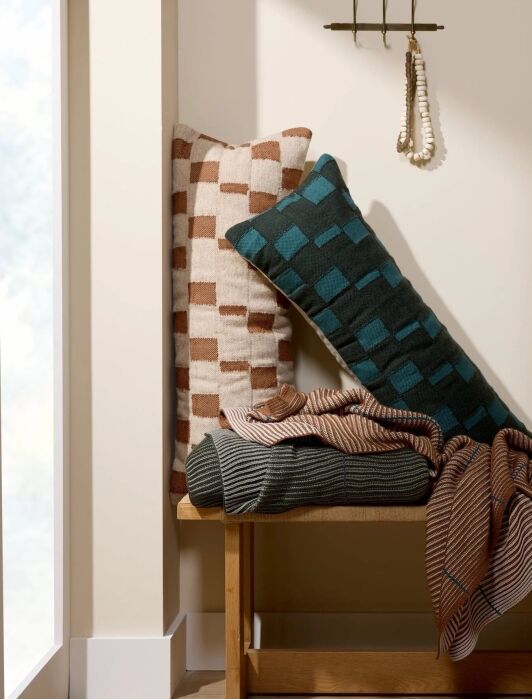Two patterned pillows and two folded blankets are arranged on a wooden bench under a small hanging rack with a beaded necklace in a neutral-colored room.