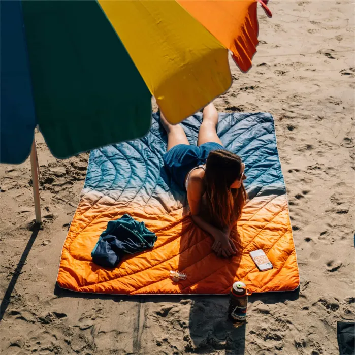 A person lies on an orange and blue blanket under a colorful beach umbrella on the sand, with a drink, sunglasses, and phone nearby.