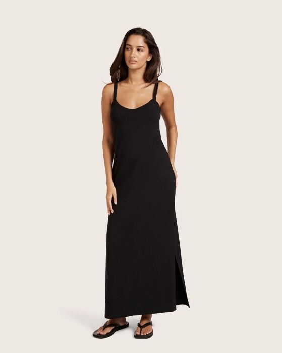A woman wearing a sleeveless black maxi dress with spaghetti straps and black sandals, standing against a plain light background.