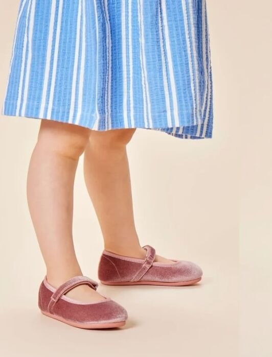 A child wearing a blue and white striped dress and pink velvet Mary Jane shoes stands on a beige background.