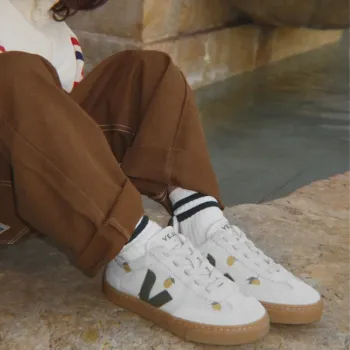 Person sitting by stone steps and water, wearing brown pants, striped socks, and white Veja sneakers with a green “V” logo and gum soles.