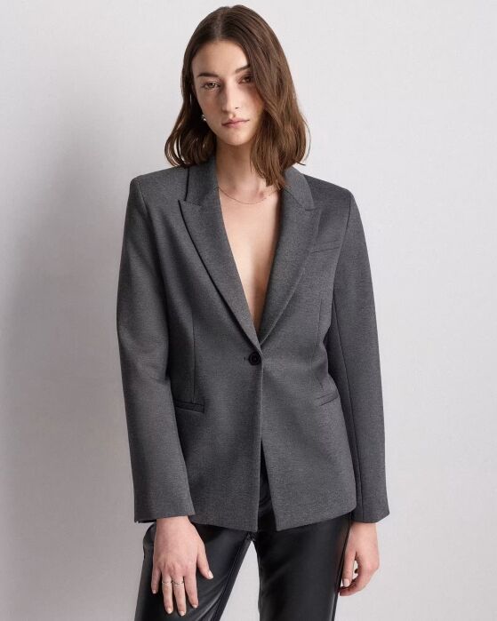 A woman with shoulder-length brown hair wears a grey blazer with no shirt underneath and black leather pants, standing against a plain light background.