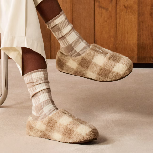 Person wearing beige checkered socks and cozy beige plaid slippers, seated on a chair with cream pants, on a light carpeted floor and wooden wall background.