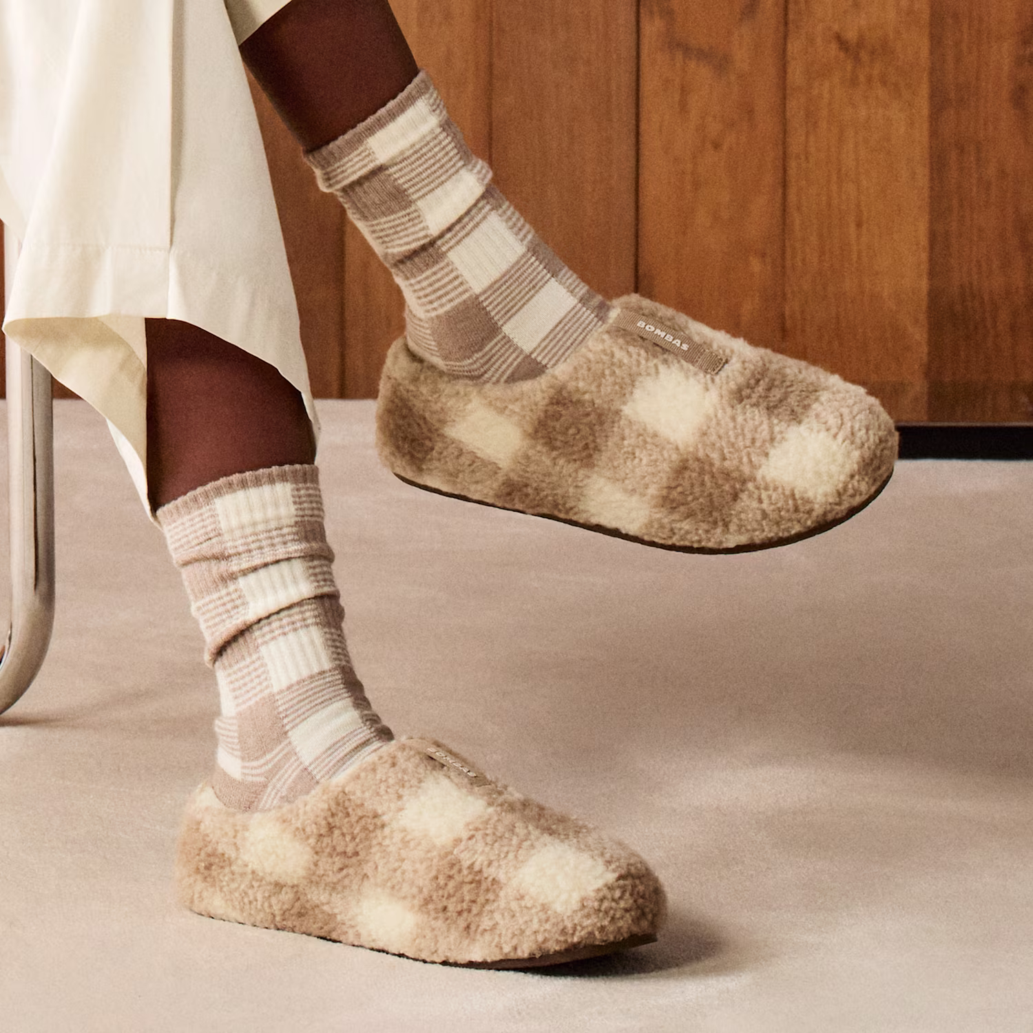 Person wearing beige checkered socks and cozy beige plaid slippers, seated on a chair with cream pants, on a light carpeted floor and wooden wall background.
