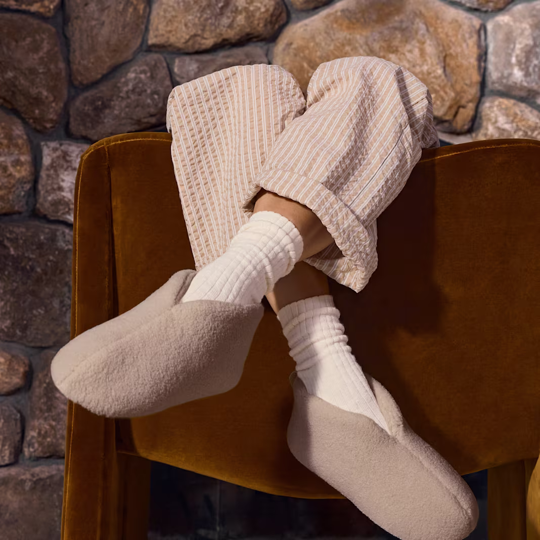 A person wearing cozy beige slippers, white socks, and striped pajama pants rests their feet on a brown chair in front of a stone wall.
