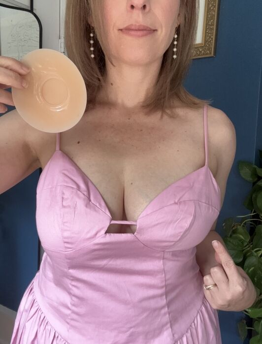 A woman in a pink dress holds a silicone breast prosthesis in one hand and gestures with her other hand, standing indoors.
