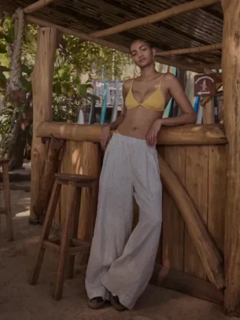A person wearing a yellow bikini top and loose gray pants leans against a wooden outdoor bar with stools, surrounded by tropical plants and surfboards.