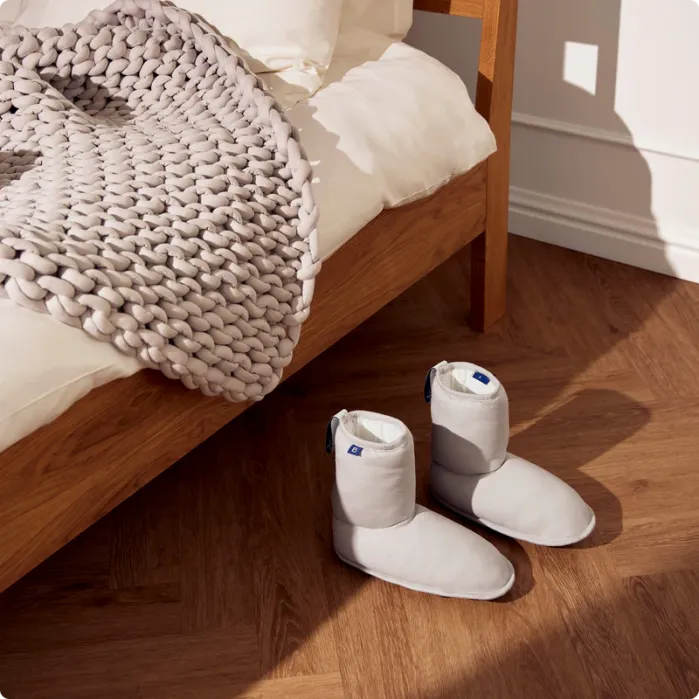 A pair of cozy slippers rest on a wooden floor beside a bed adorned with a chunky knit blanket and crisp white bedding.