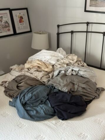 A pile of assorted colored bedsheets and linens is spread across a neatly made bed in a bedroom with framed pictures on the wall.