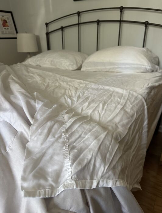 A bed with white, slightly wrinkled sheets and two pillows sits against a wall with a metal headboard. A lamp and framed picture are visible in the background.