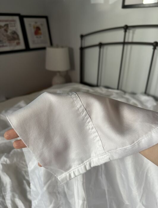 A hand holds up the cuff of a white satin long-sleeve shirt or pajama top against a bed and lamp in a softly lit bedroom.