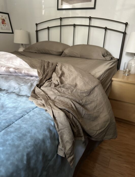 A made bed with taupe sheets and pillows, a metal headboard, and a beige nightstand. A blanket is partially pulled back, exposing a blue comforter underneath.