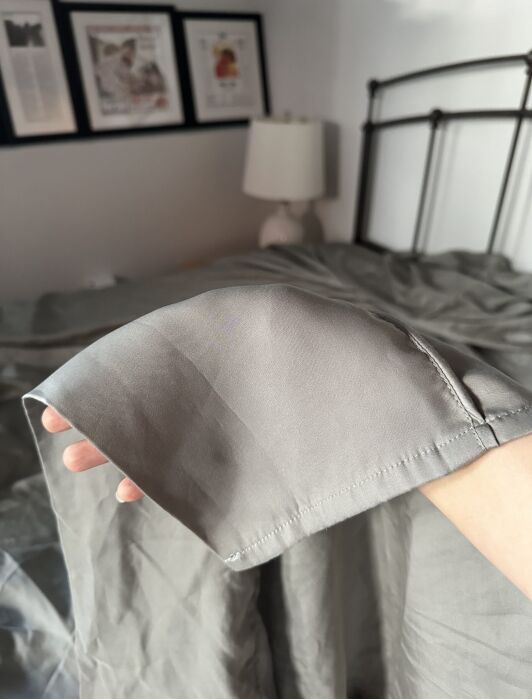 A hand holds the edge of a gray satin sheet in a bedroom with a bed, white lamp, and framed pictures on the wall in the background.