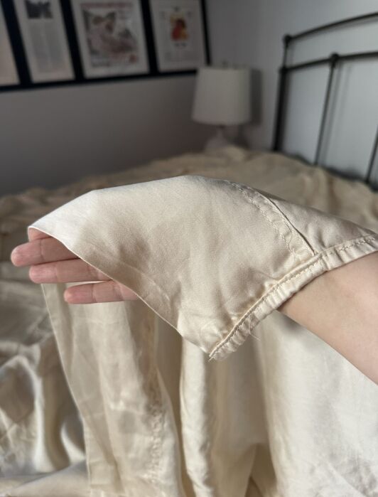 A hand holds up the corner of a light beige silk sheet in a bedroom with framed pictures on the wall in the background.