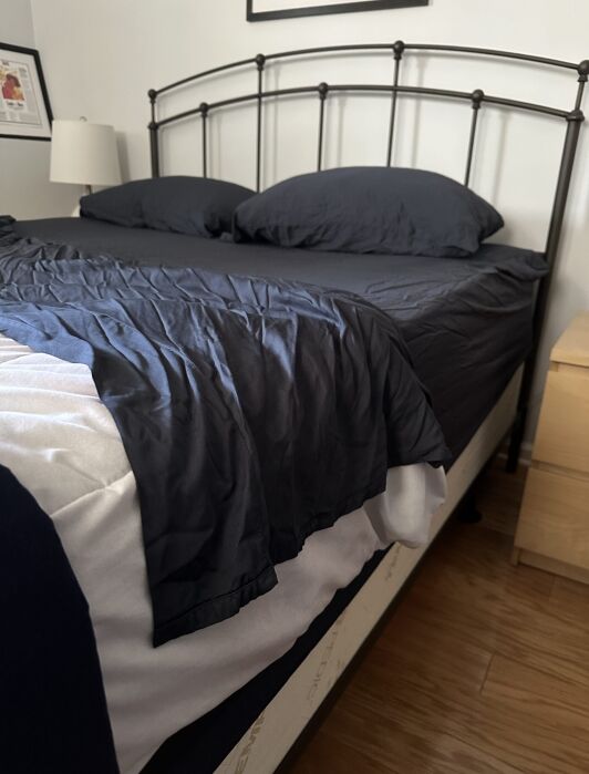 A neatly made bed with dark gray sheets and two pillows, next to a light wood nightstand, set on a wooden floor in a minimally decorated bedroom.