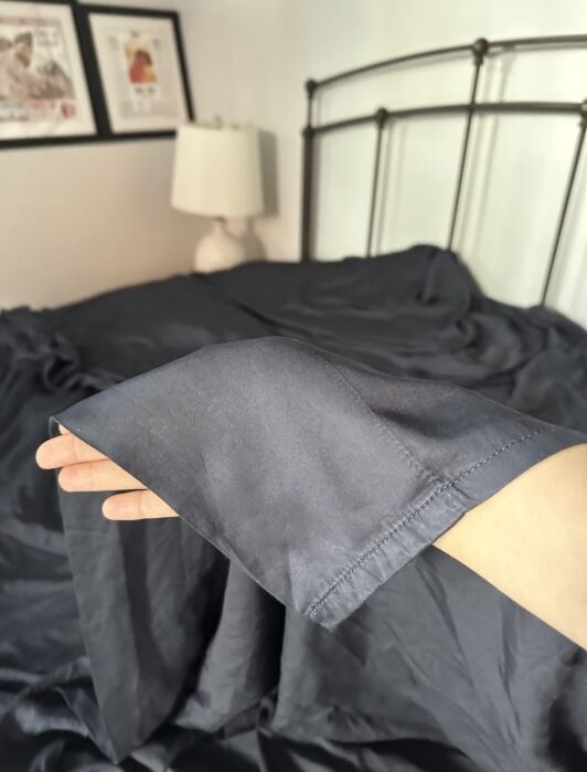 A hand holds the corner of a dark gray bedsheet in front of an unmade bed with a metal headboard, lamp, and framed art on the wall.