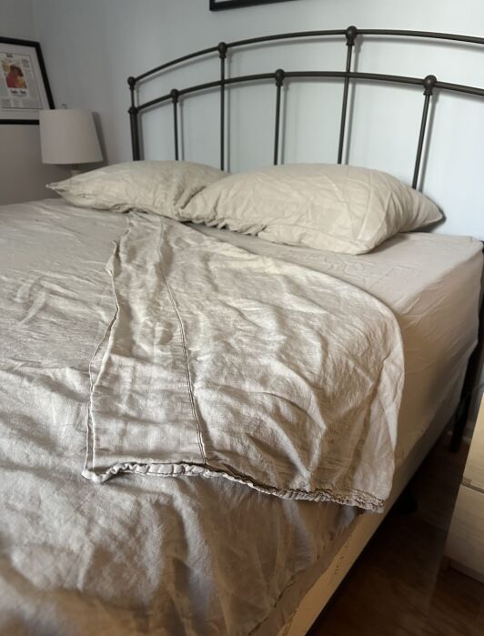 Unmade bed with beige sheets and two pillows, one pillowcase wrinkled, against a metal headboard. A lamp and framed photo are on the bedside table.