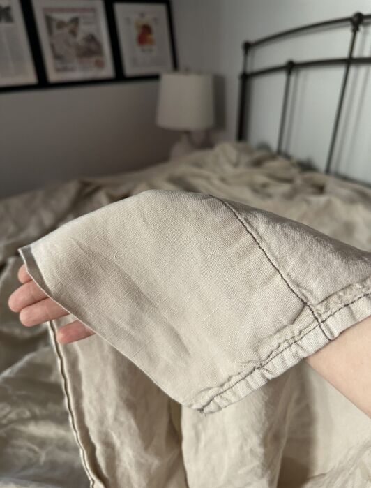 A hand holds up the corner of a beige linen blanket in a bedroom with framed pictures, a lamp, and a metal bedframe visible in the background.