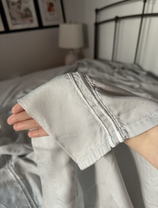 A hand holds up the corner of a light gray bedsheet in a bedroom with a lamp, metal headboard, and framed pictures on the wall.