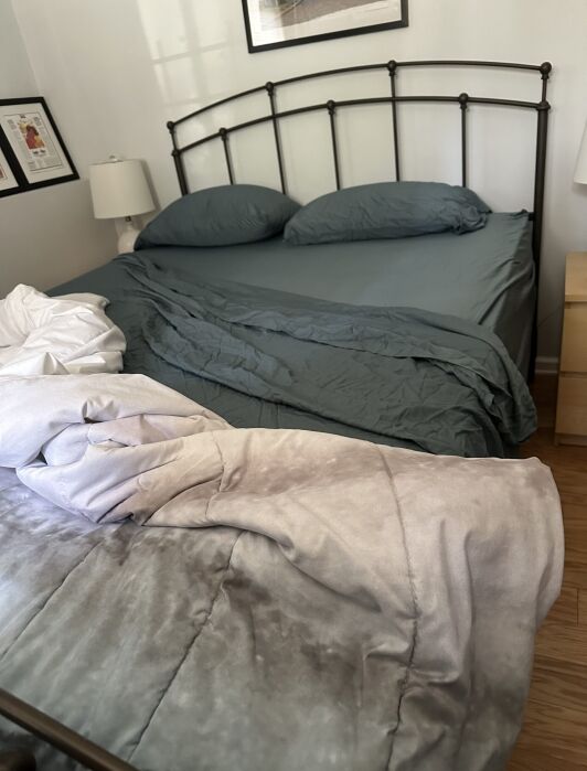 A bed with dark green sheets and two pillows, partially unmade, with a light gray blanket draped messily over the edge and onto the floor.