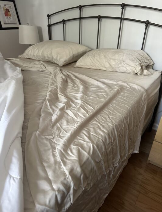 A queen-sized bed with beige sheets and two pillows, partially unmade, sits next to a nightstand and a lamp, on a wooden floor.