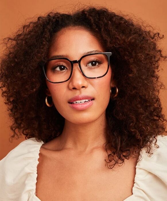 Woman with curly hair, wearing large black glasses, gold hoop earrings, and a white top, looking slightly up and to the right against a plain brown background.