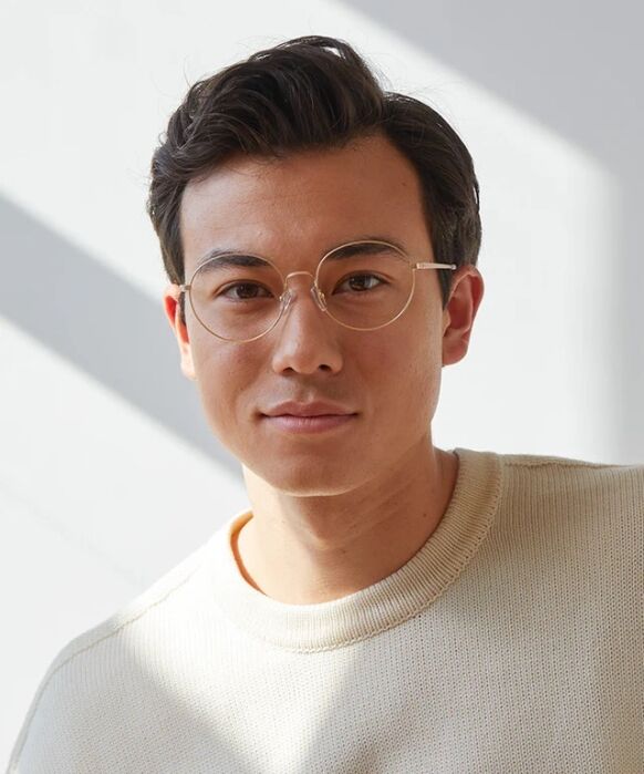 A person with short dark hair and round glasses wears a cream-colored sweater, standing in soft natural light against a plain background.