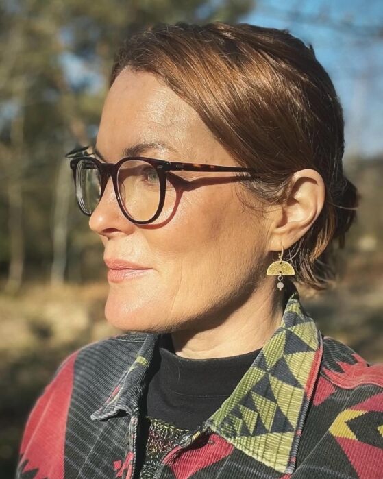 Woman with short brown hair and glasses looks to the left, wearing a patterned jacket and gold earrings, with sunlight illuminating her face outdoors.