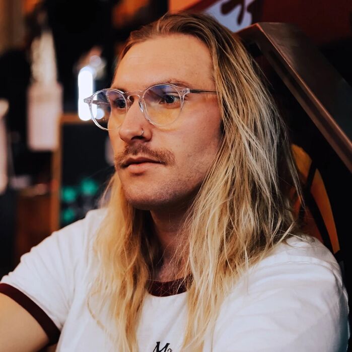 A person with long blonde hair, glasses, and a mustache sits indoors wearing a white T-shirt with dark trim, looking thoughtfully to the side.