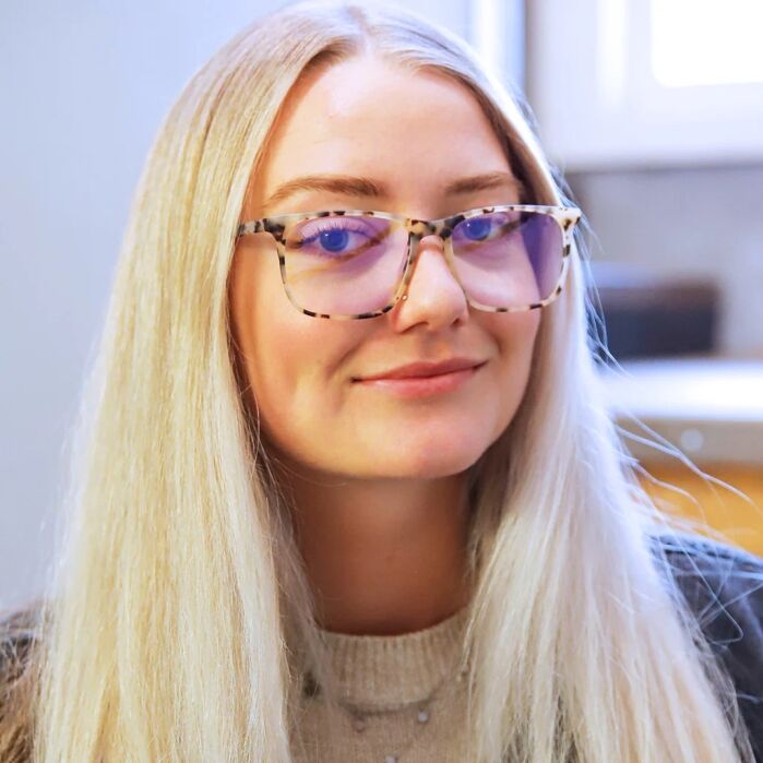 A woman with long blonde hair and tortoiseshell glasses looks at the camera and smiles slightly. She is indoors with a blurred background.