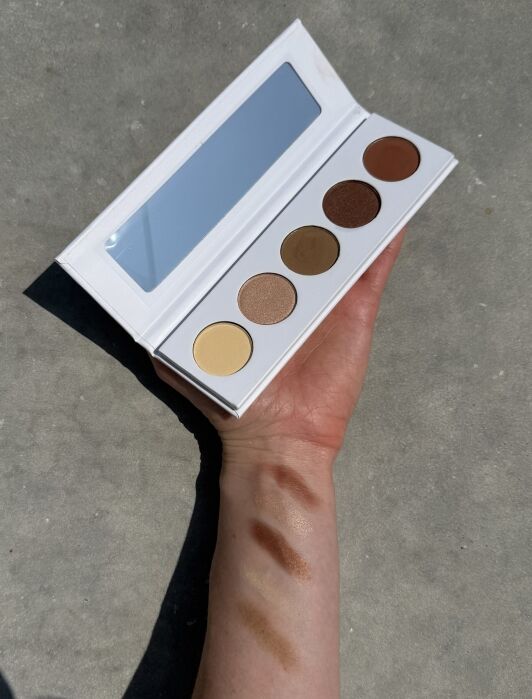 A hand holds an open eyeshadow palette with five shades; swatches of the colors are visible on the forearm.