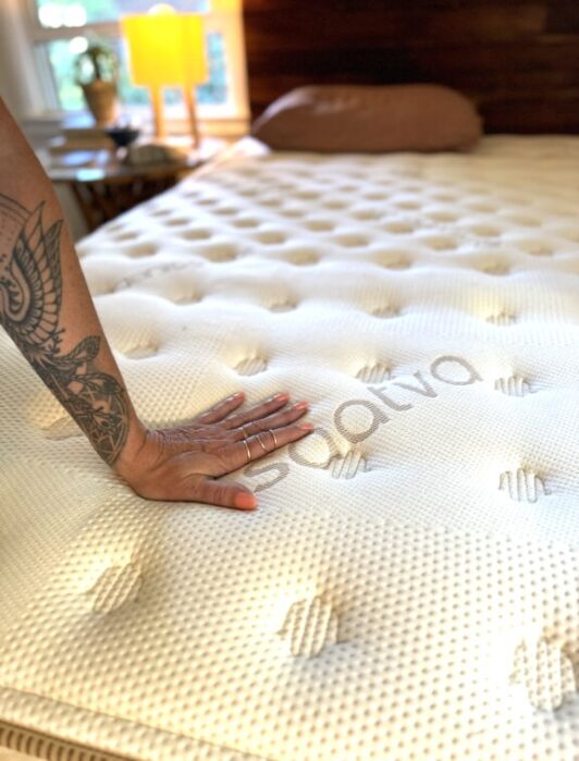 A tattooed hand presses down on a Saatva mattress in a sunlit bedroom, showing the mattress’s texture and firmness.