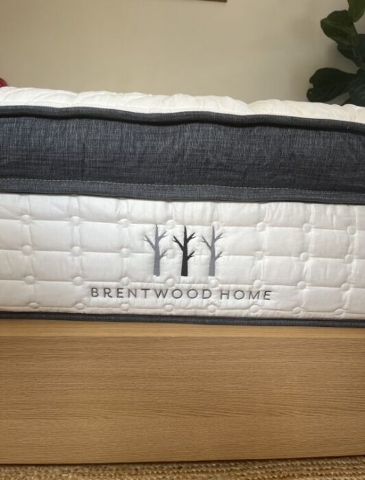 Side view of a Brentwood Home mattress on a wooden bed frame, showing quilted fabric and the brand’s tree logo embroidered in gray.