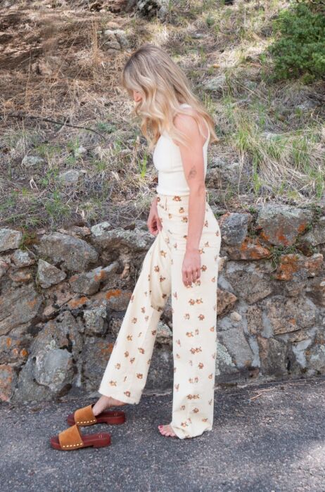 A woman with wavy blonde hair, wearing a white top and cream-colored floral pants, stands barefoot on a paved path near a stone wall. Brown sandals are on the ground beside her.
