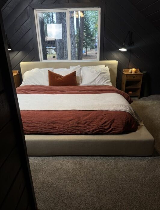 A neatly made bed with white and rust-colored bedding, flanked by two wooden nightstands, sits below a window overlooking trees in a carpeted bedroom.