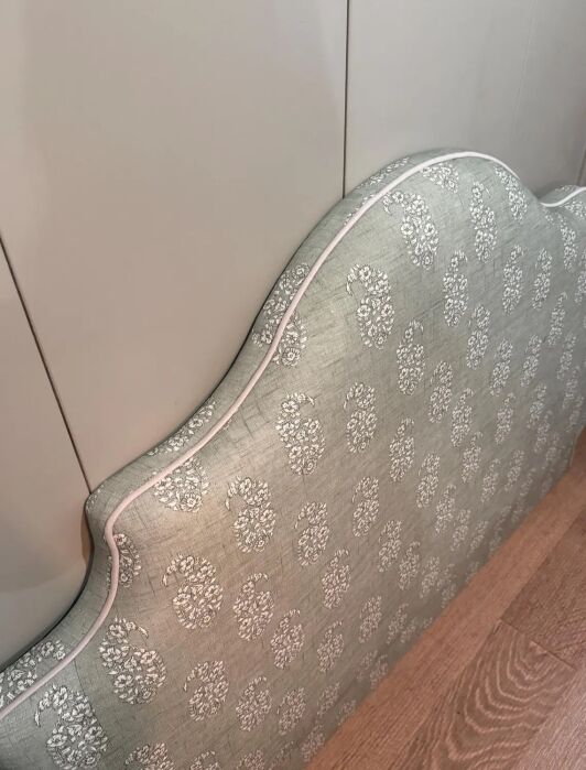Upholstered headboard with a light blue and white floral pattern, white piping, leaning against a gray wall on a wooden floor.