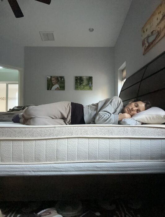 A woman lies on her side asleep on an organic mattress in a dimly lit bedroom, with two framed photos and a painting visible on the wall behind her.