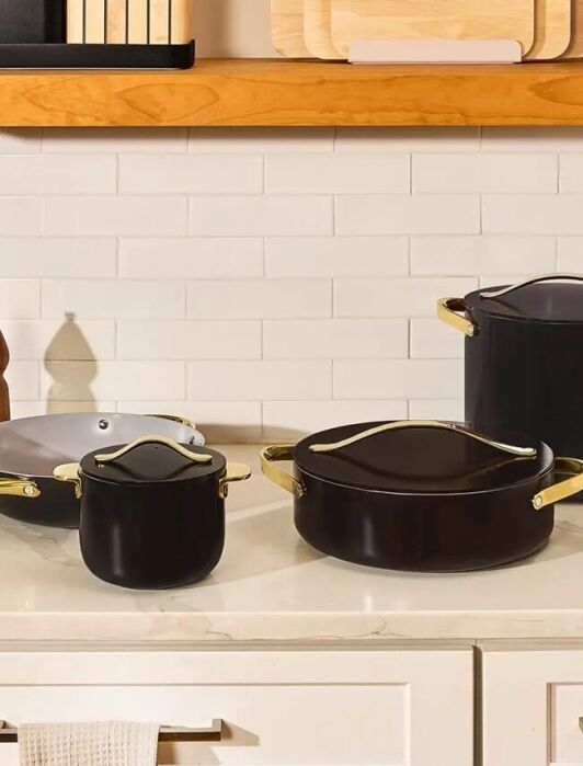 A set of black cookware with gold handles, including a frying pan, saucepan, Dutch oven, and stockpot, arranged on a kitchen countertop against a white tile backsplash.