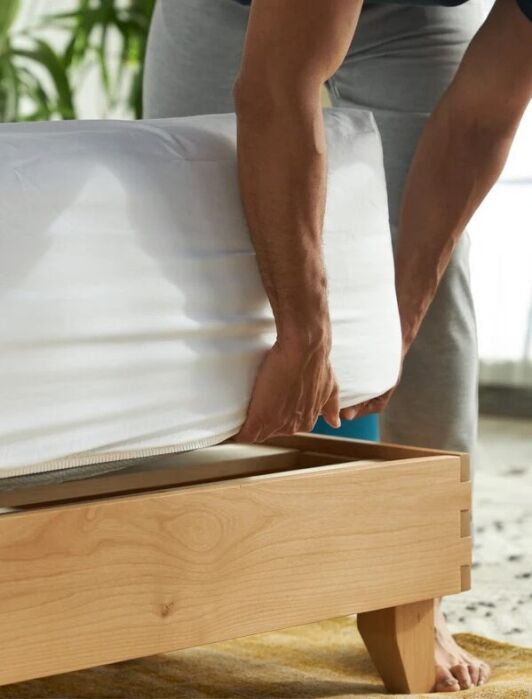 A person pulls up a white mattress cover on a wooden bed frame, with cooling sheets and green plants visible in the background.