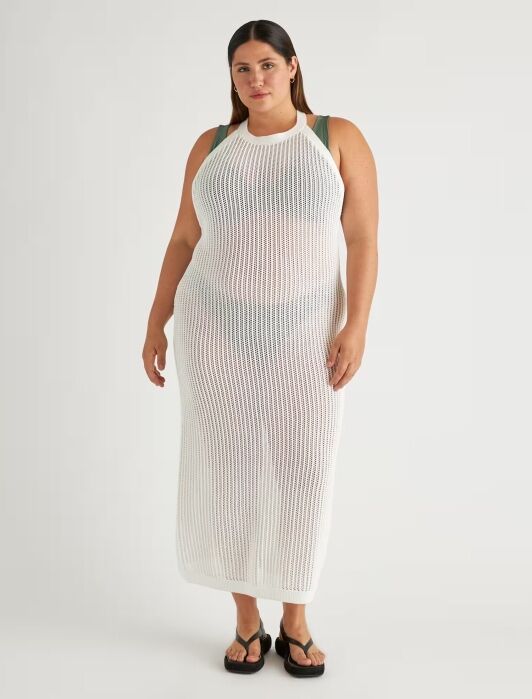 A woman stands facing forward wearing a sleeveless, long, white knit cover-up dress over a green bikini and black sandals against a plain background.