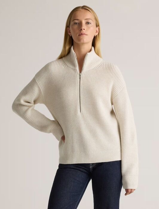 A person wearing a cream-colored, ribbed quarter-zip sweater and dark blue jeans stands against a plain light background.
