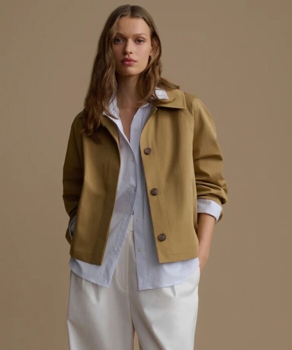 A woman stands against a beige background, wearing a tan jacket over a white button-up shirt and white pants, with hands in her pockets.