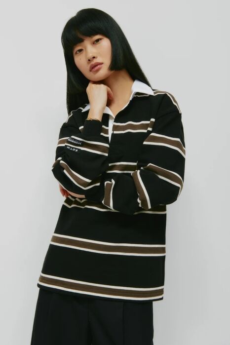 A person with straight black hair wears a black, brown, and white striped long-sleeve rugby shirt and dark pants, standing against a plain light background.