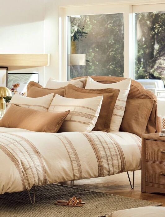 A neatly made bed with cream and tan striped bedding, several pillows, and a beige rug in a sunlit bedroom with modern decor and large window.