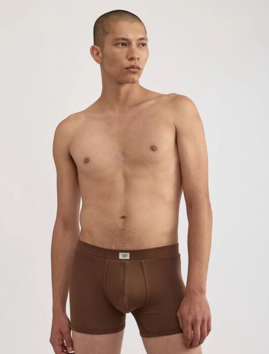 A man with a buzz cut stands against a plain background, wearing only brown boxer briefs and looking to the side.