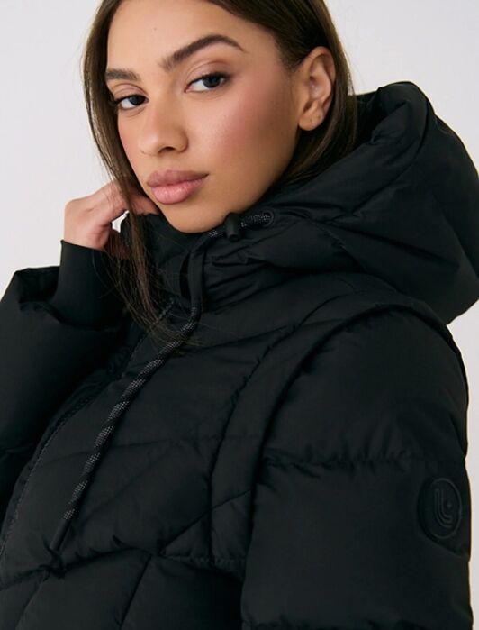 A woman wearing a black puffer jacket with a hood looks at the camera, her hand resting near her neck.