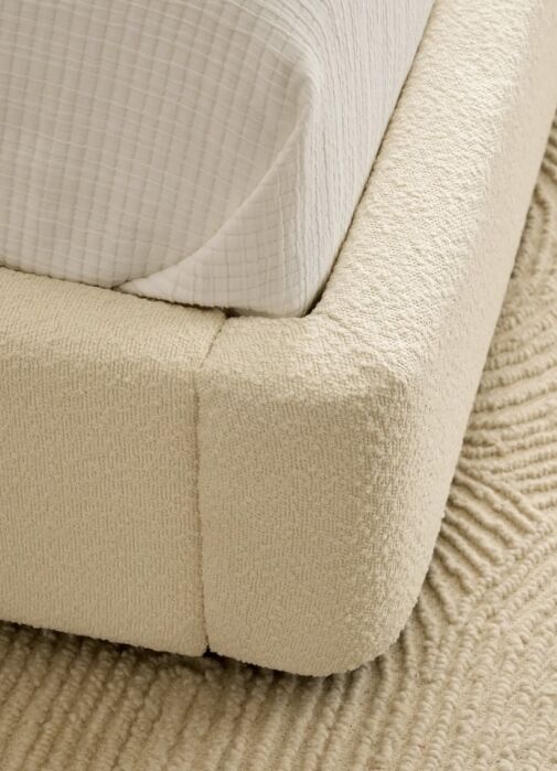 Close-up of a cream-colored upholstered bed frame corner with a white textured blanket, resting on a beige textured rug.